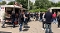 Menschen stehen auf einem gepflasterten Hof vor einem Pizza-Imbisswagen und einem weiteren Verkaufswagen unter blauem Himmel mit Wolken