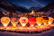 Sieben beleuchtete Heißluftballons stehen in einer verschneiten Berglandschaft vor einem Dorf mit einer Kirche unter einem sternenklaren Nachthimmel, umgeben von zahlreichen Menschen.