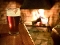 Ein Glas Bier steht auf einem Holztisch vor einem Kamin mit brennendem Feuer