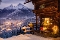 Verschneite Berghütte mit beleuchteter Fensterdekoration und Blick auf ein beleuchtetes Tal vor schneebedeckten Bergen im Abendlicht