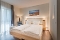 Hell eingerichtetes Schlafzimmer mit Doppelbett, zwei weißen Kissen, Nachttischlampen, Stehlampe, kleinem runden Tisch mit Wasserflasche und Glas sowie einem Landschaftsbild über dem Bett