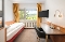 Kleines Hotelzimmer mit Einzelbett, Schreibtisch, schwarzem Stuhl, Wandfernseher und Fenster mit gelben Vorhängen und Blick ins Grüne.